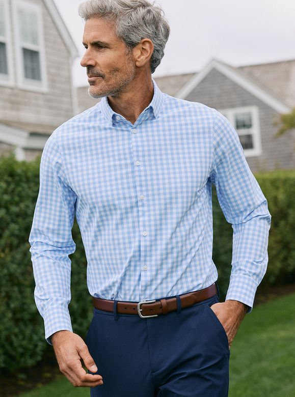 Gray-haired man walking across a lawn in a light blue gingham dress shirt, navy pants and a brown belt