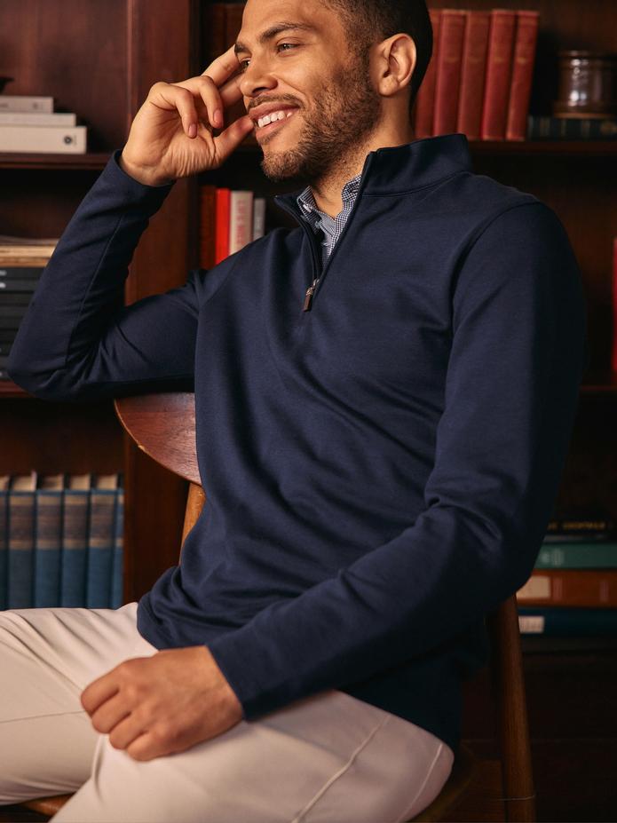 A smiling man sitting in a wooden chair indoors wearing a navy blue quarter-zip pullover over a patterned collared shirt and light beige trousers.