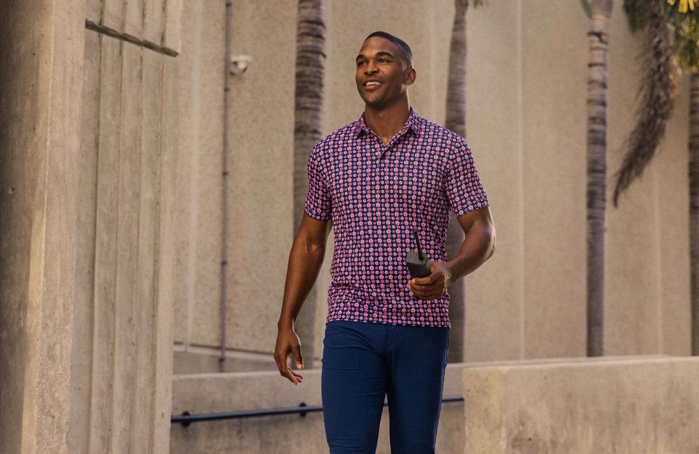A smiling model, walking along the outside of a concrete building, wearing a red and blue patterned Mizzen+Main short sleeve shirt, while holding a vintage cell phone.