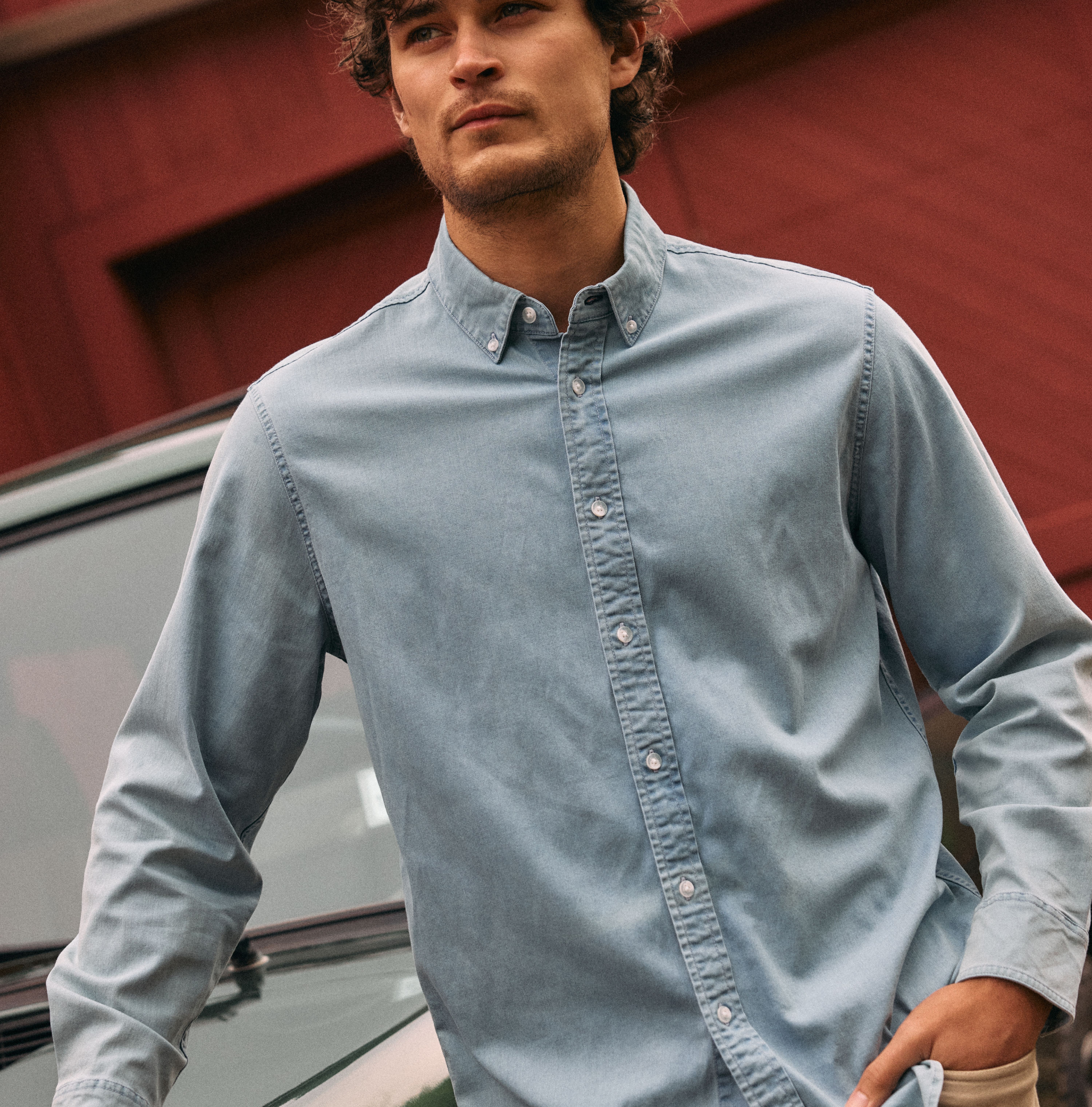 A man with curly hair leaning against a green vehicle in front of a red building, wearing a light blue denim-look button-down shirt and beige pants.