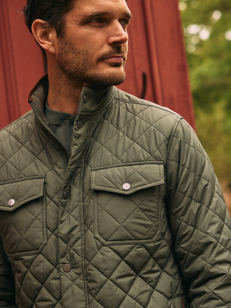 A close-up portrait of a man wearing an olive green quilted jacket standing in front of a red building wall.