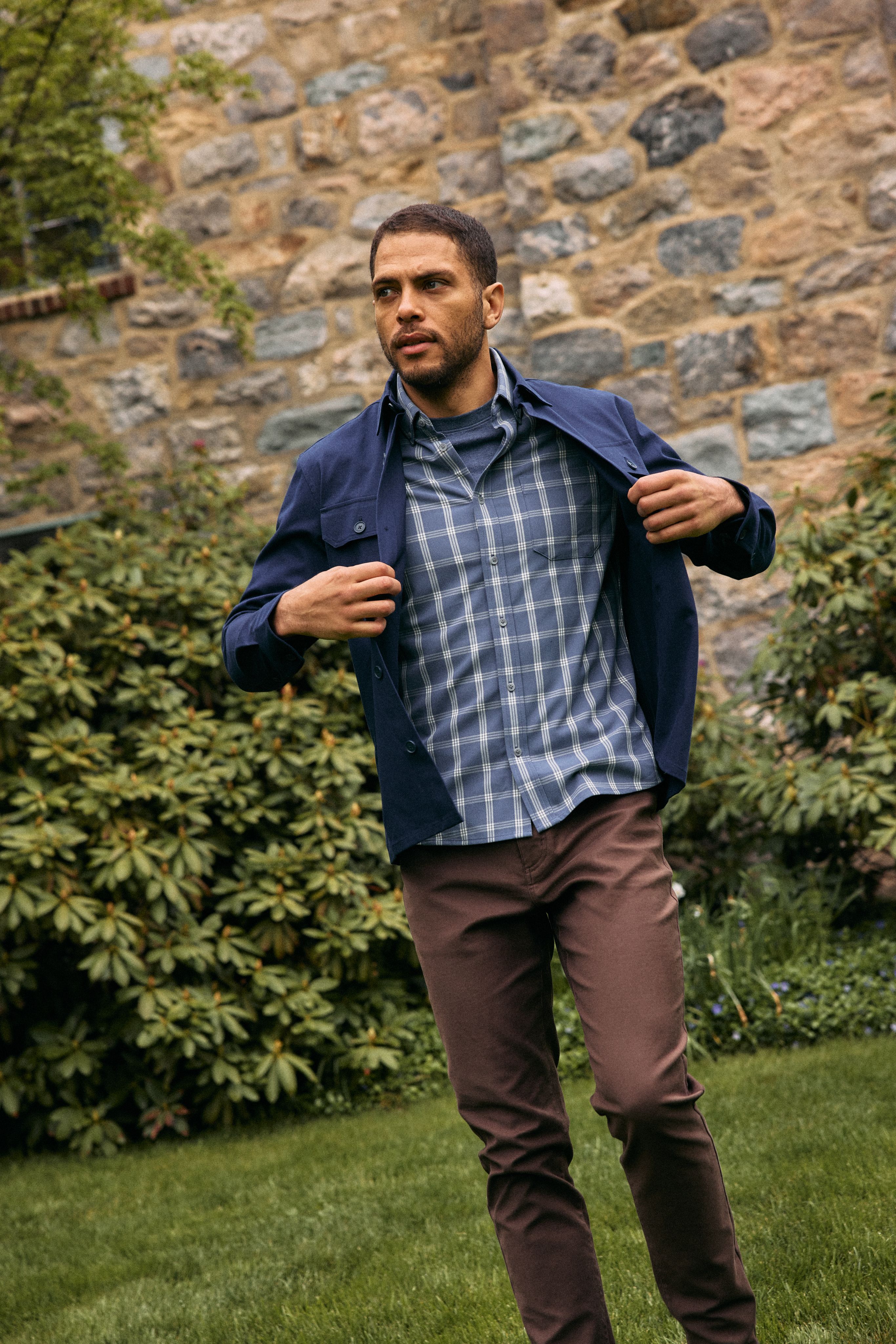 Man pulling on a navy shirt jacket over a blue plaid shirt with brown pants in front of a stone wall