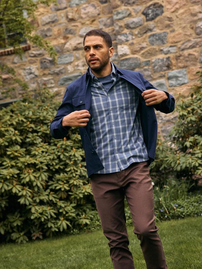 Man pulling on a navy shirt jacket over a blue plaid shirt with brown pants in front of a stone wall