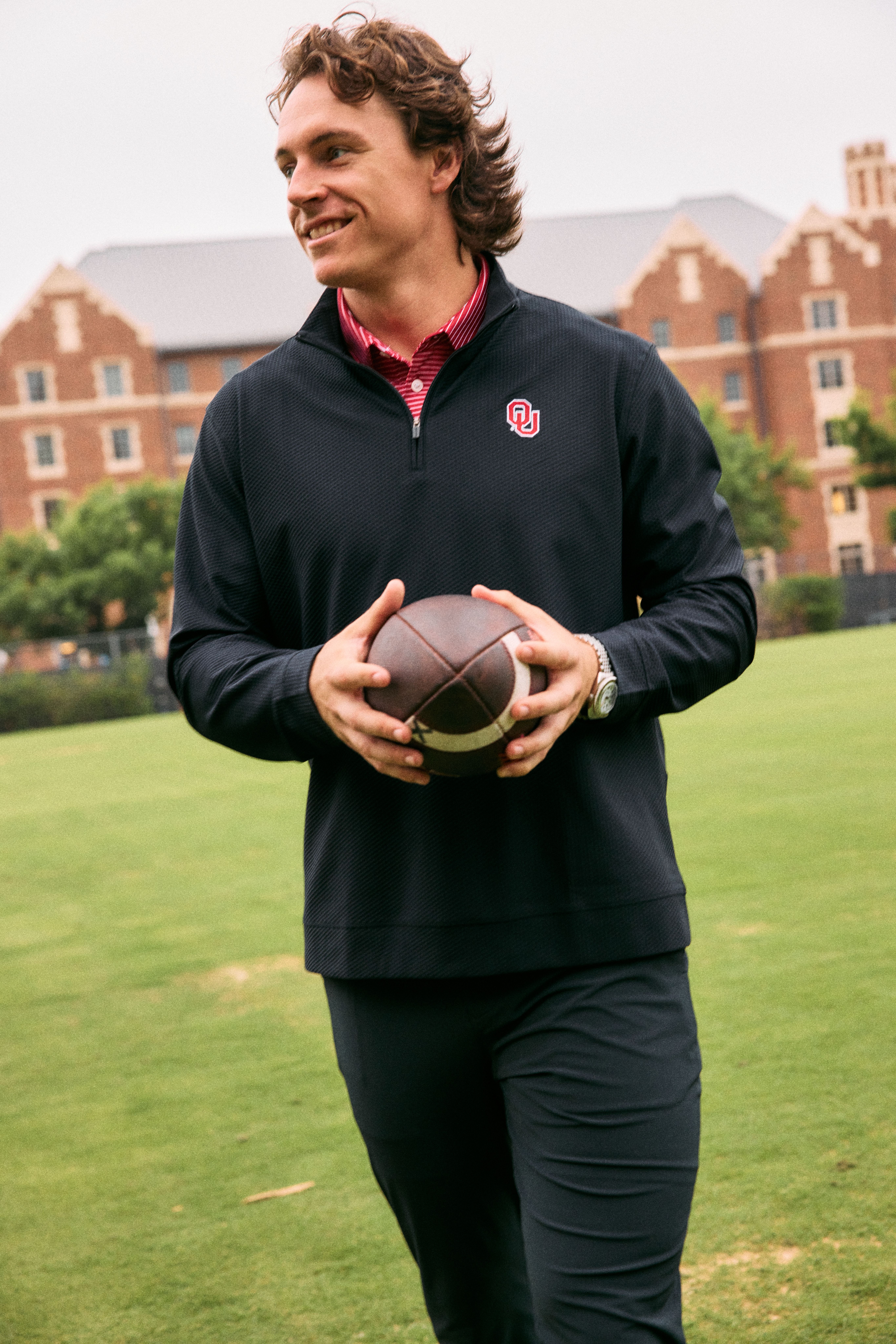 A smiling John Mateer, Oklahoma Sooners quarterback, holding a football while walking across a grassy campus field. He is wearing a dark quarter-zip pullover featuring the University of Oklahoma logo over a red striped collar.