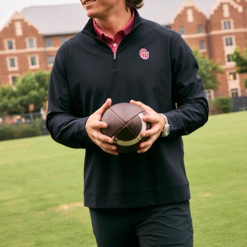 A smiling John Mateer, Oklahoma Sooners quarterback, holding a football while walking across a grassy campus field. He is wearing a dark quarter-zip pullover featuring the University of Oklahoma logo over a red striped collar.