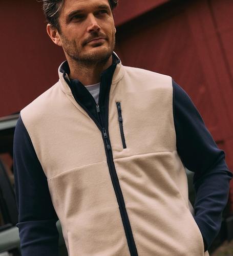 A man wearing a beige fleece vest with a dark blue collar and zipper trim over a dark long-sleeve shirt.