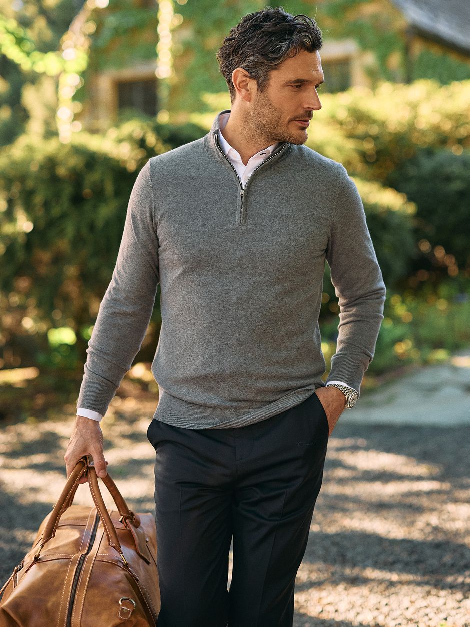 A man in a grey quarter-zip sweater, white collared shirt, and black trousers carrying a brown leather duffel bag outdoors.