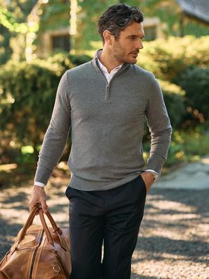 A man in a grey quarter-zip sweater, white collared shirt, and black trousers carrying a brown leather duffel bag outdoors.
