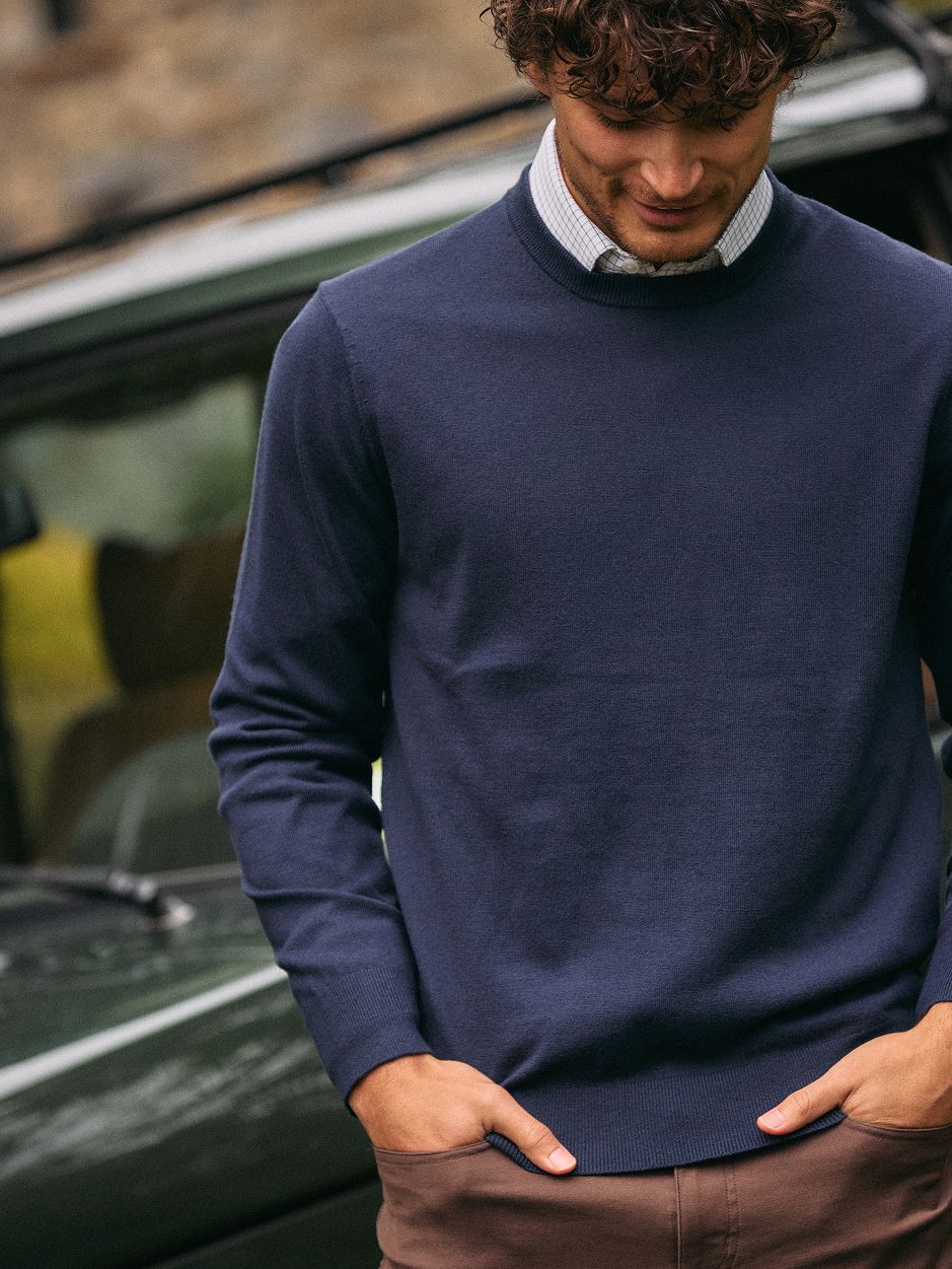 A man in a dark navy crewneck sweater, white collared shirt, and brown trousers standing in front of a vehicle.