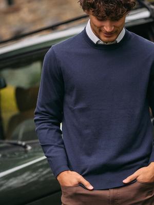 A man in a dark navy crewneck sweater, white collared shirt, and brown trousers standing in front of a vehicle.