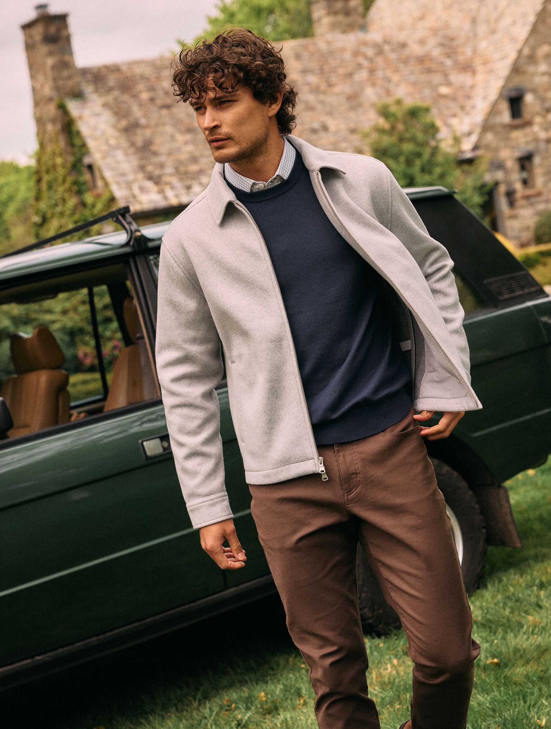 A man in a light grey full-zip jacket, navy sweater, collared shirt, and brown pants standing near a vintage green SUV.