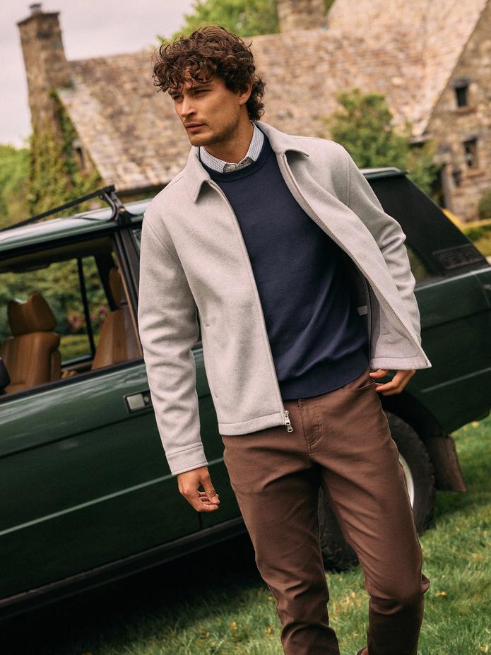 A man in a light grey full-zip jacket, navy sweater, collared shirt, and brown pants standing near a vintage green SUV.