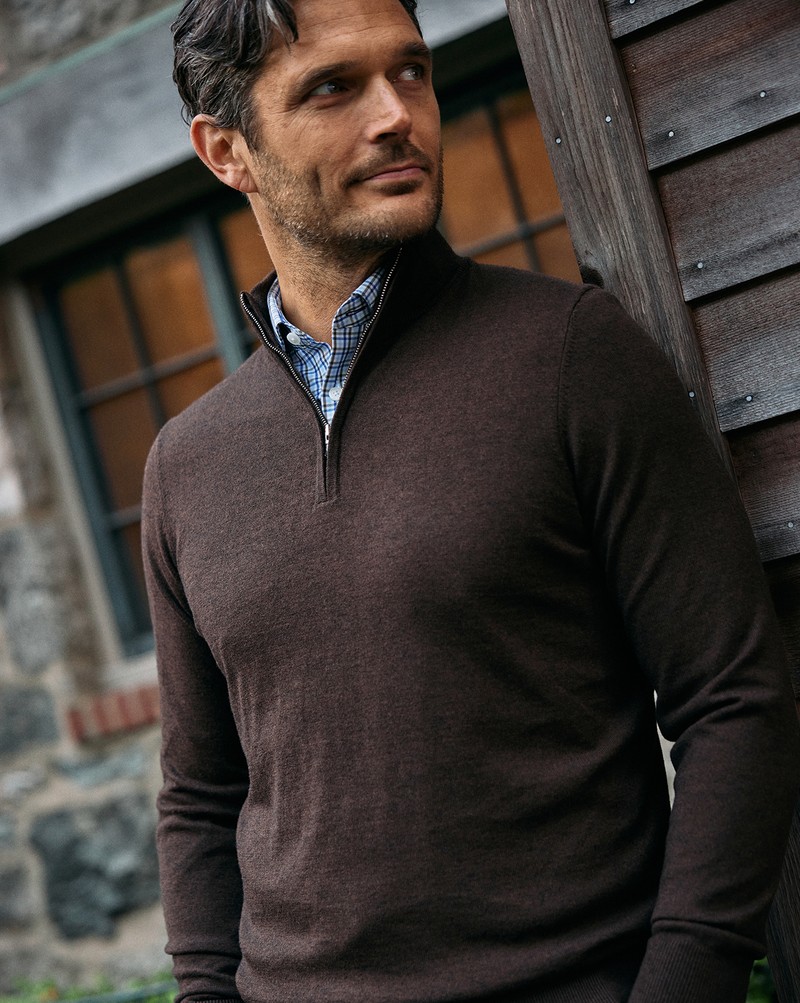 Man wearing a brown Heritage Quarter Zip Sweater over a blue check button-down, leaning against a wooden building.
