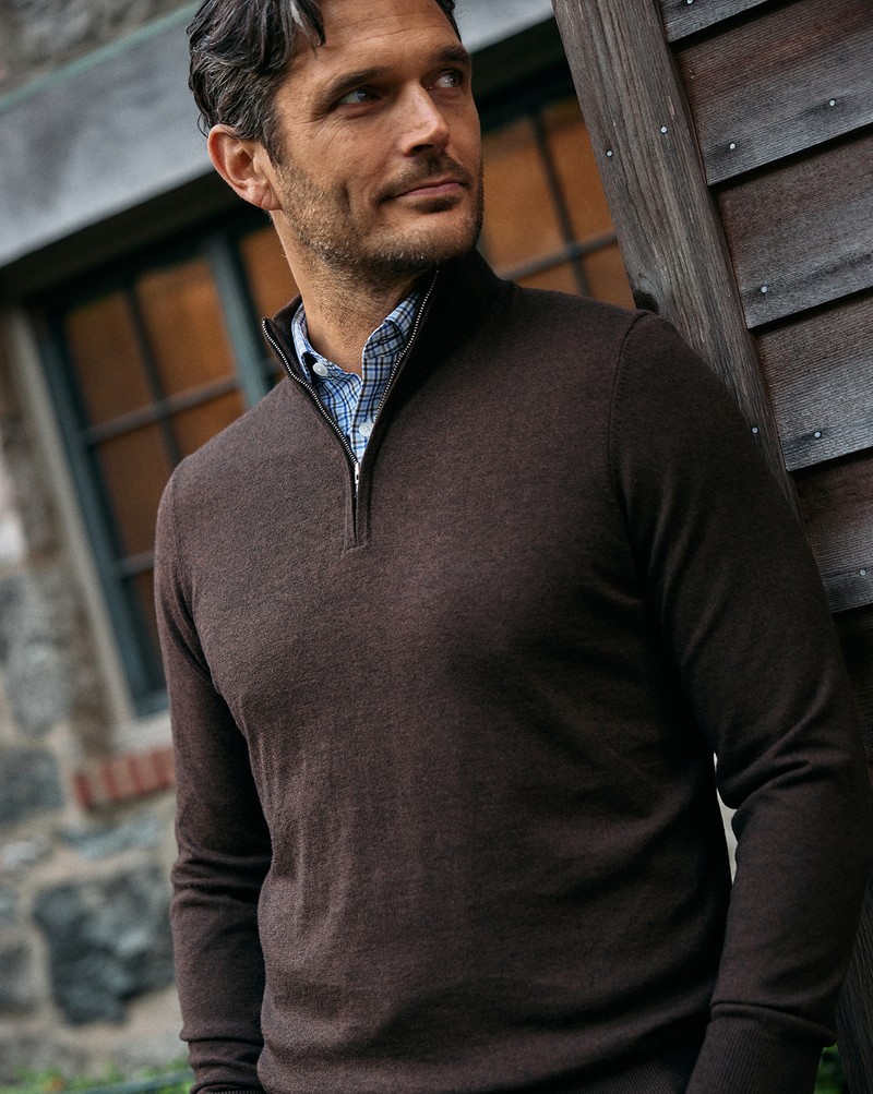 Man wearing a brown Heritage Quarter Zip Sweater over a blue check button-down, leaning against a wooden building.