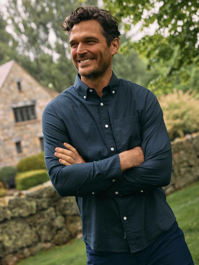 A smiling man standing outdoors with arms crossed, wearing a dark blue grid-pattern button-down shirt against a stone building background.