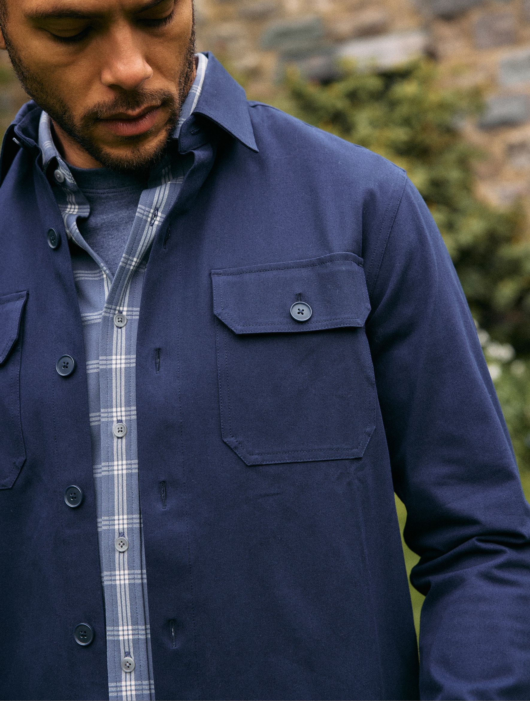 A close-up shot of a man wearing a dark navy button-front overshirt with flap chest pockets layered over a blue plaid button-down shirt.