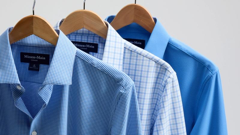 Three button-down shirts on wooden hangers, showing blue gingham, blue plaid, and solid blue Mizzen+Main Leeward styles.