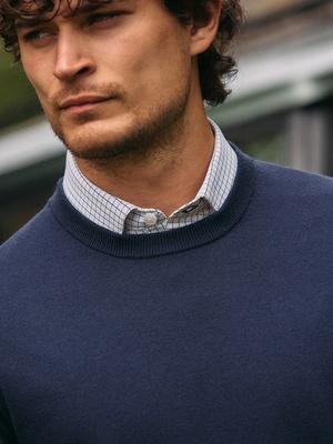 A close-up portrait shot of a man wearing a dark navy crewneck sweater layered over a white micro-grid collared shirt.