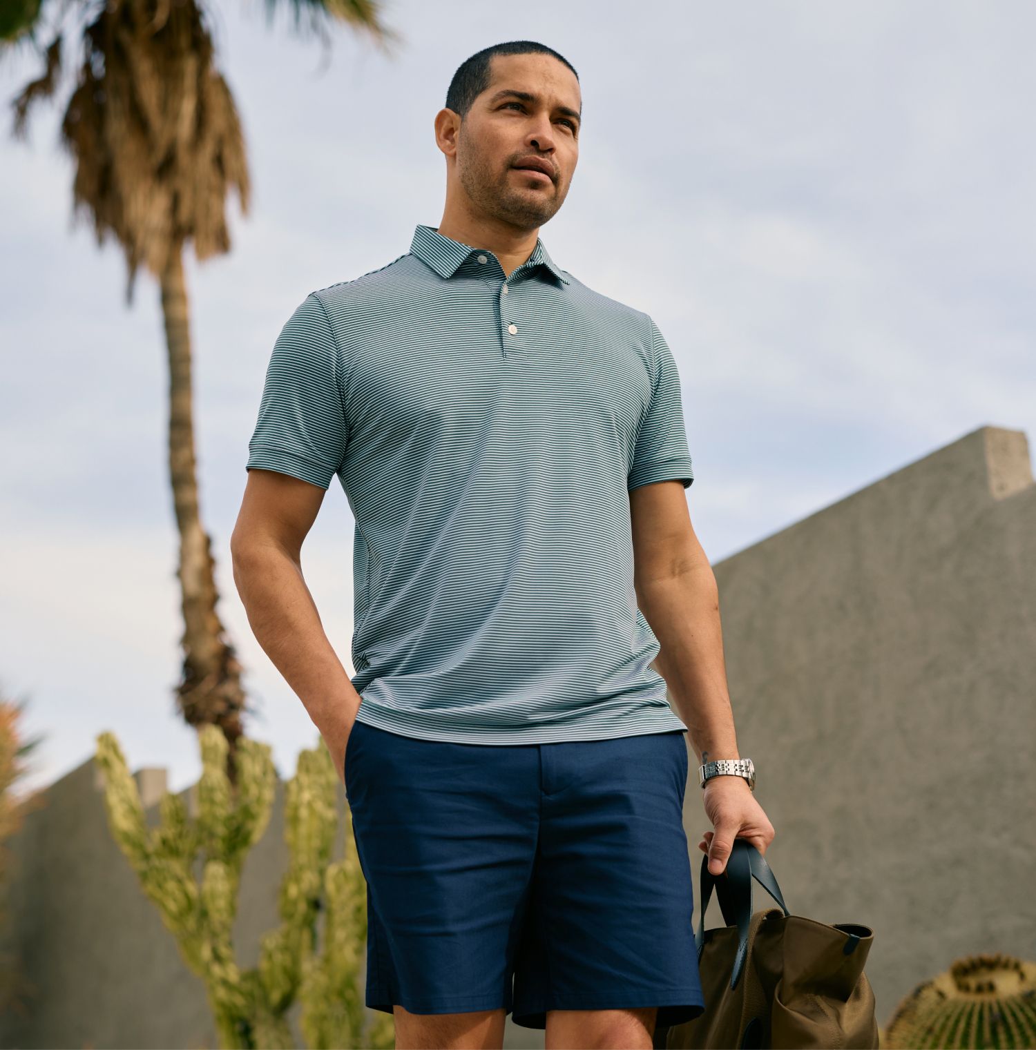 Man in a green and white striped polo shirt and navy shorts standing outdoors near a palm tree and cactus, holding a bag.