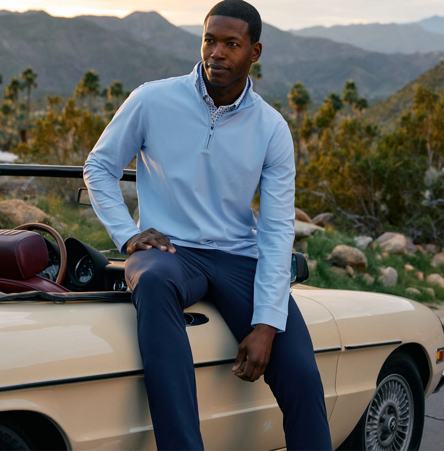 Man in a light blue quarter-zip pullover and navy pants sitting on the hood of a cream vintage convertible, desert mountains and palm trees in the background.