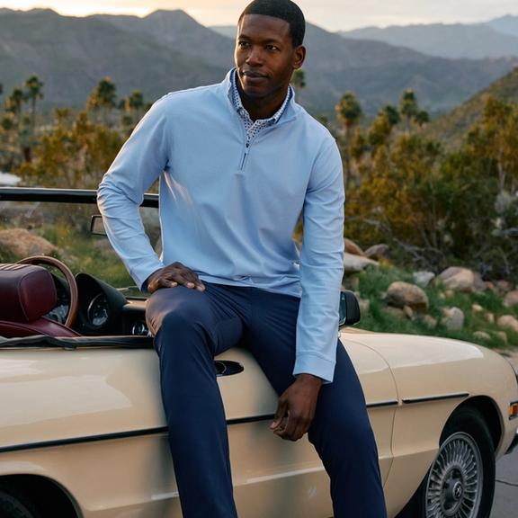 Man in a light blue quarter-zip pullover and navy pants sitting on the hood of a cream vintage convertible, desert mountains and palm trees in the background.