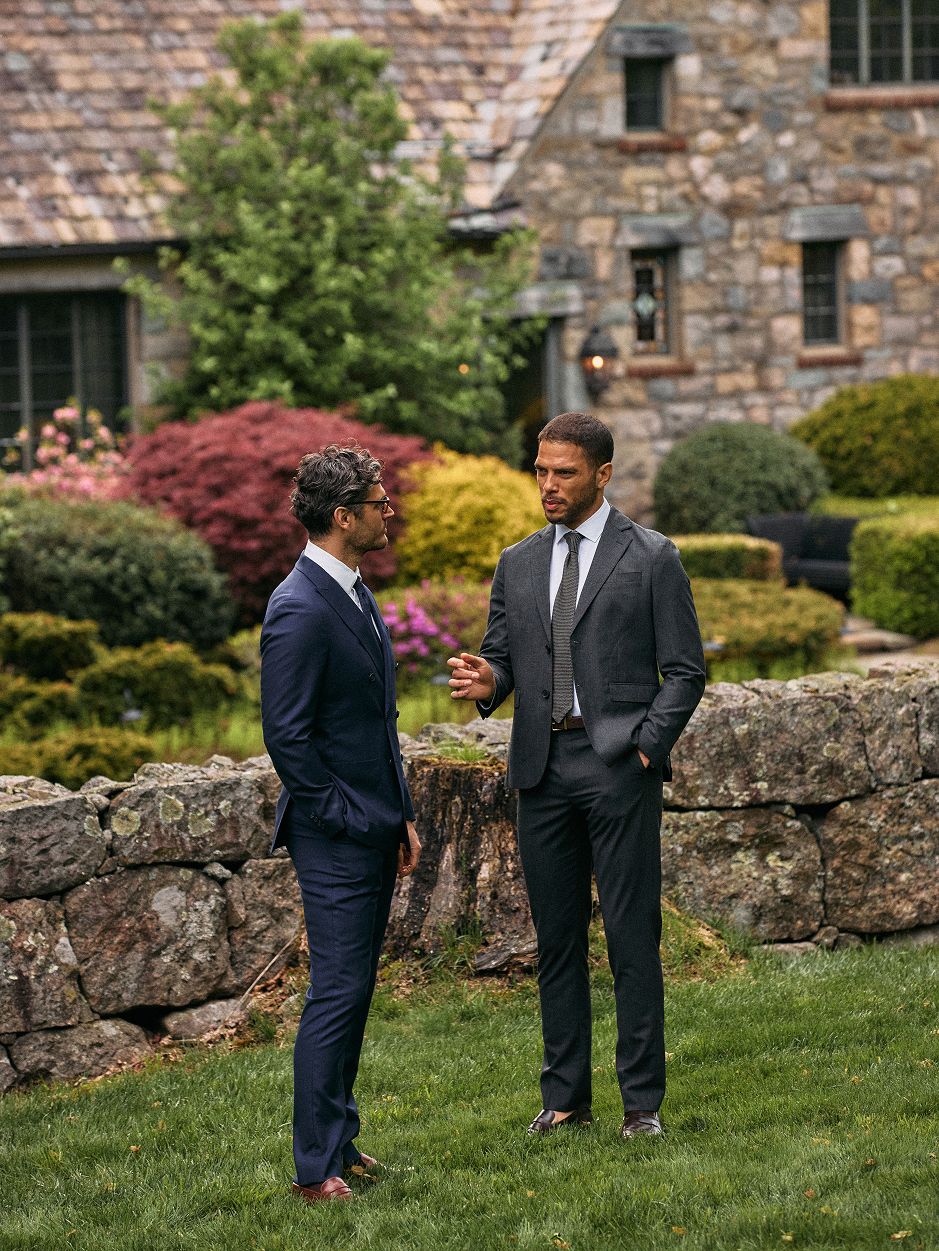 Two men in suits standing outdoors in a garden area near a stone wall, conversing with a stone house in the background.