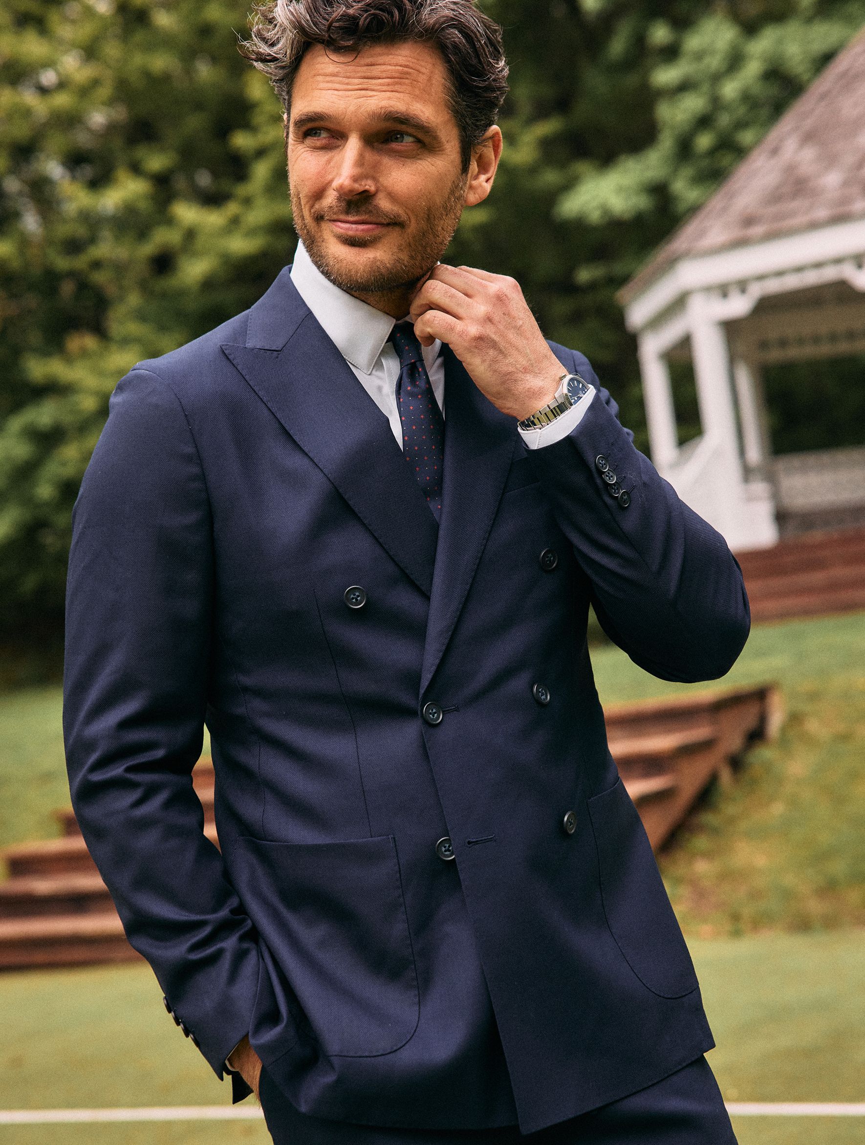 A man adjusting his collar outdoors, dressed in a double-breasted navy suit, white shirt, and patterned tie with a white gazebo in the background.