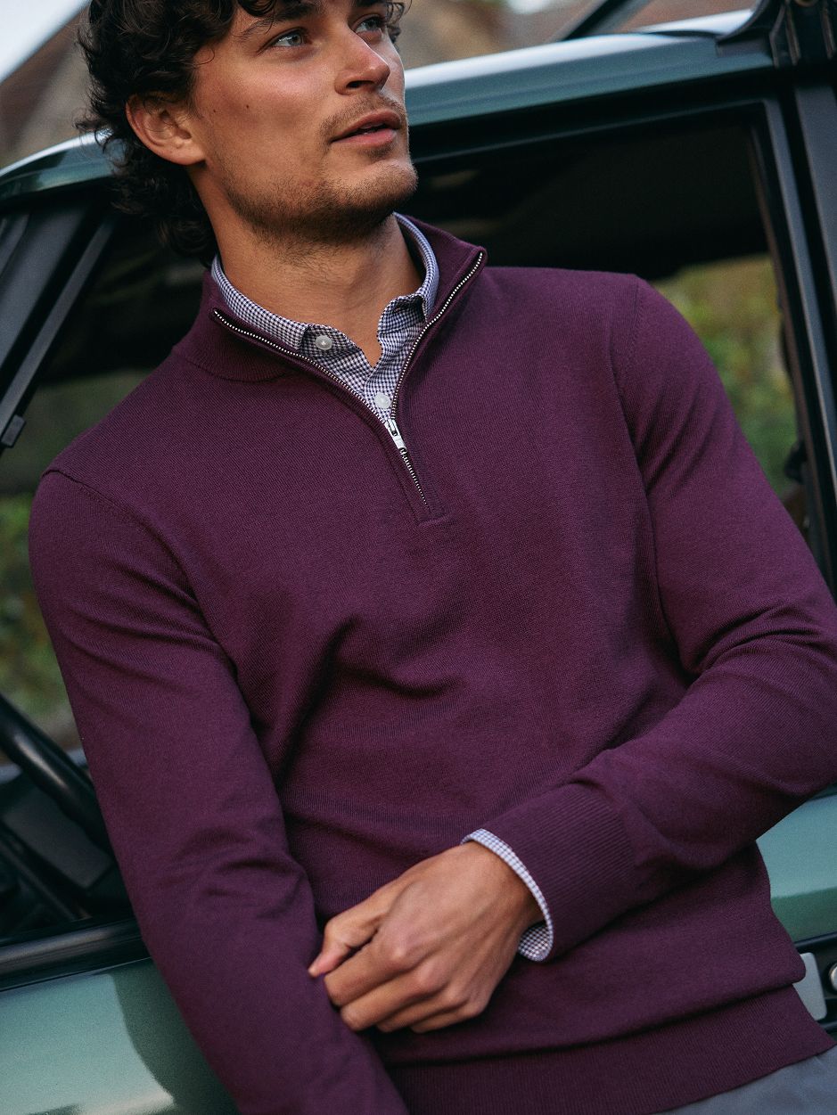 A man in a deep purple quarter-zip sweater layered over a patterned collared shirt, leaning against a vehicle.