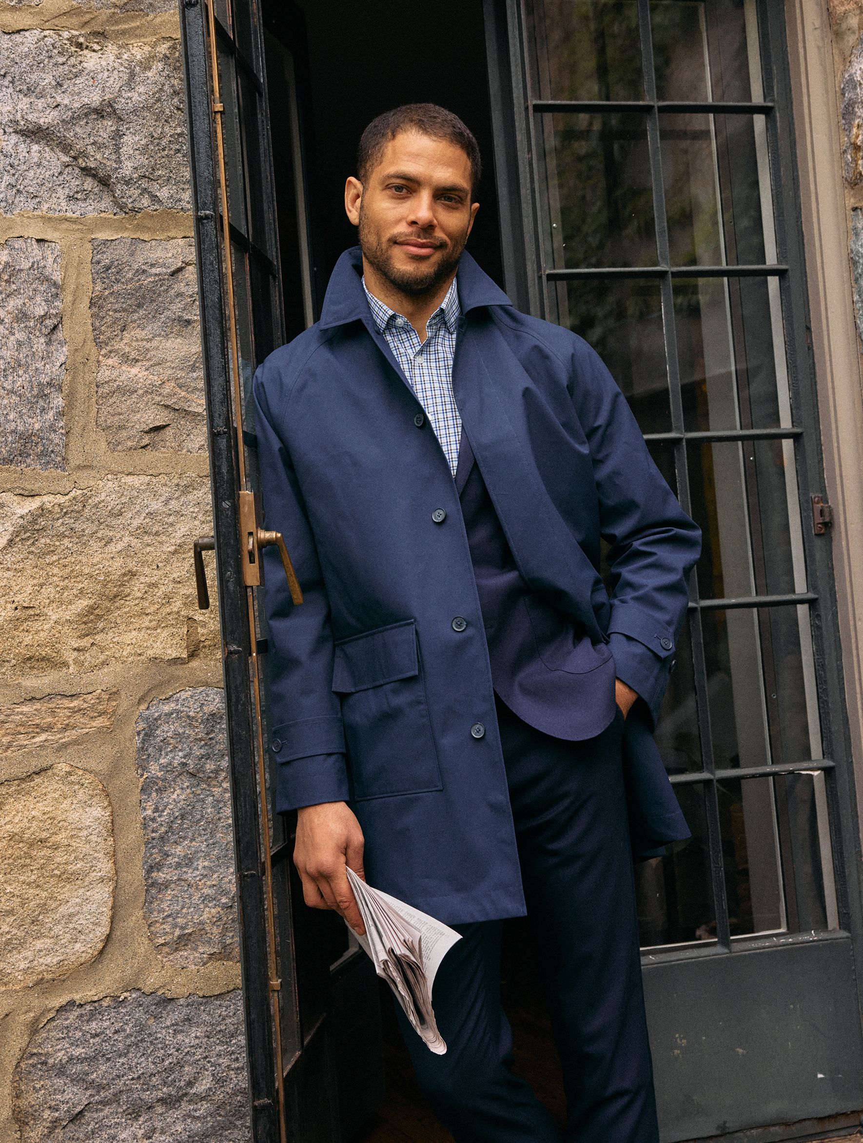 A man holding a folded newspaper standing at an open glass-and-stone doorway, wearing a navy coat over a patterned collared shirt and dark trousers.