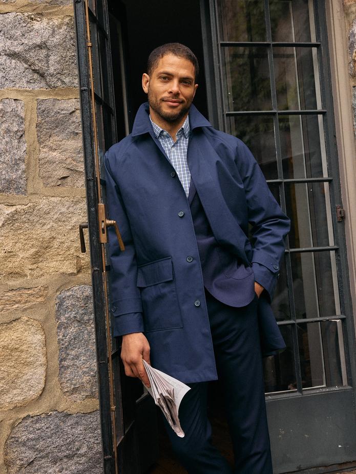 A man holding a folded newspaper standing at an open glass-and-stone doorway, wearing a navy coat over a patterned collared shirt and dark trousers.