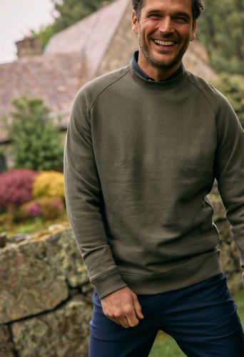 Smiling man in an olive green crewneck sweatshirt over a collared shirt, with navy pants, standing beside a stone wall in a cottage garden