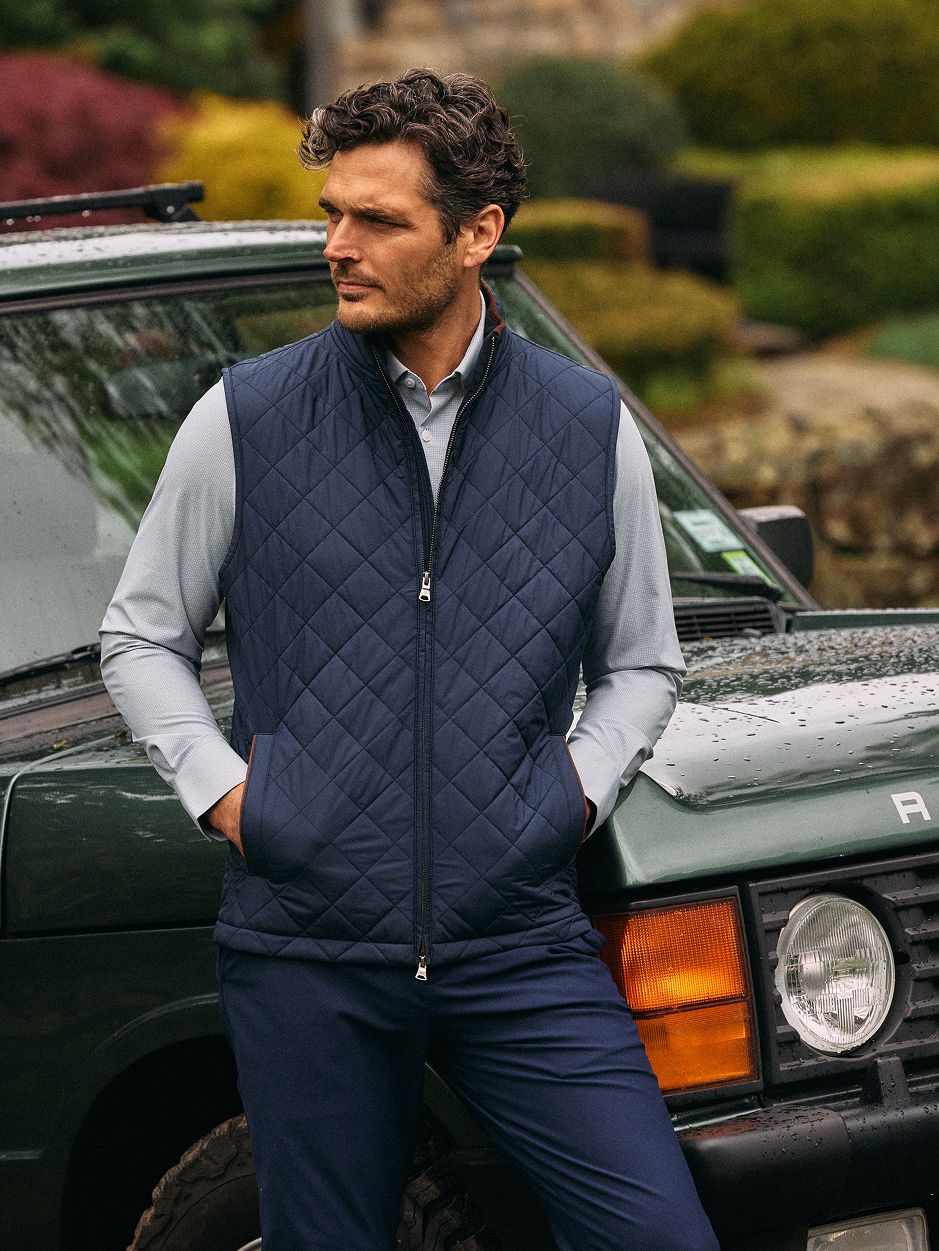 A man with hands in his pockets wearing a navy blue quilted vest over a grey long-sleeve shirt and navy pants, leaning against a green SUV.