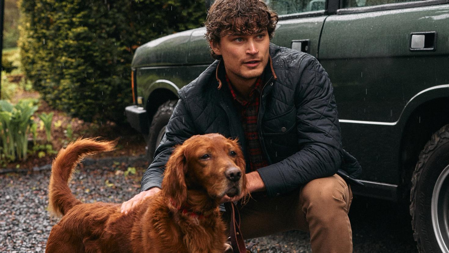 A man in a black quilted jacket crouching down next to an Irish Setter dog outside near a green SUV in the rain.