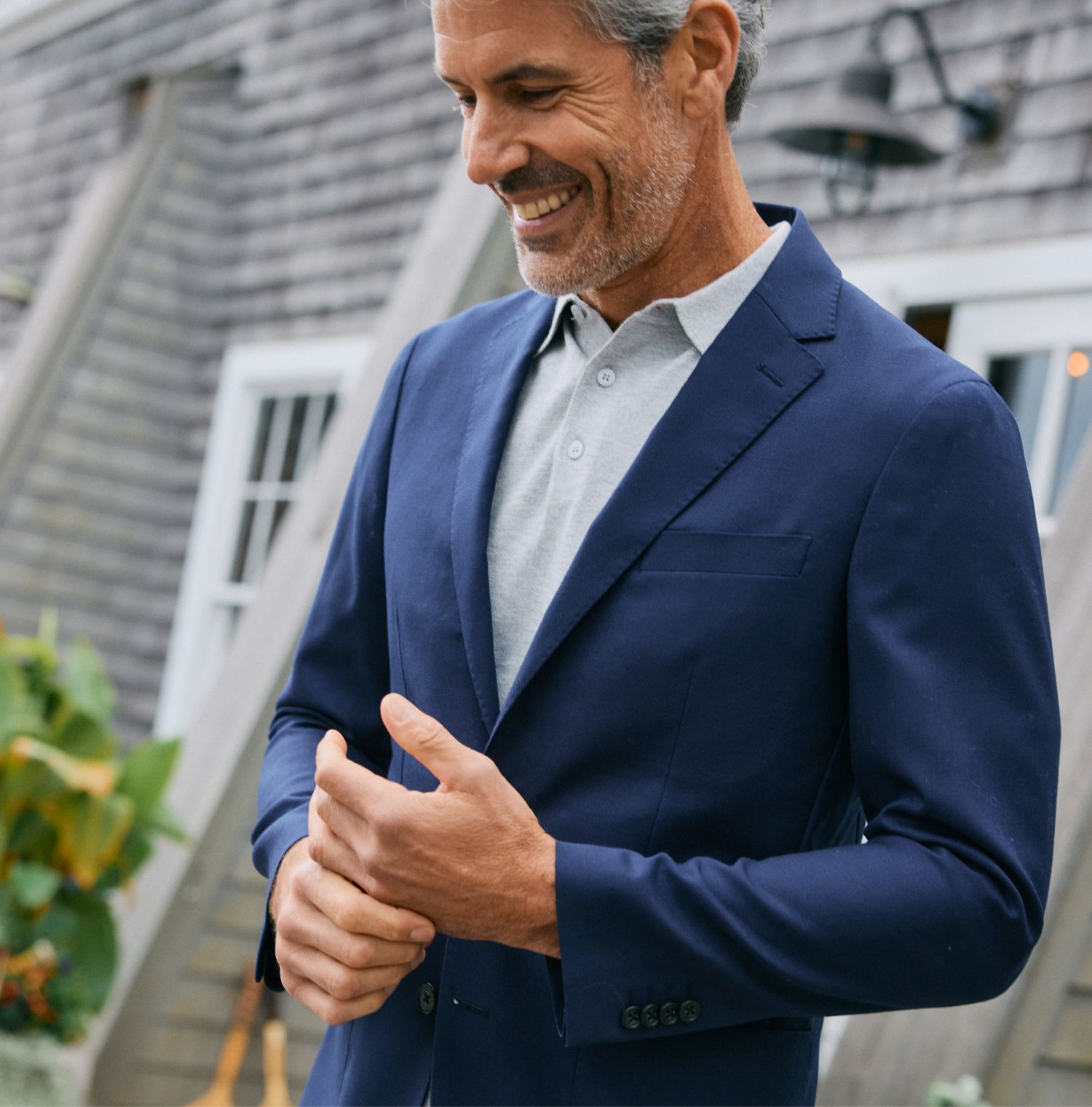 Close-up of a smiling man in a navy blazer layered over a light gray polo shirt, standing outside a shingled house.