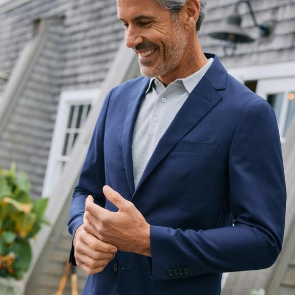 Close-up of a smiling man in a navy blazer layered over a light gray polo shirt, standing outside a shingled house.