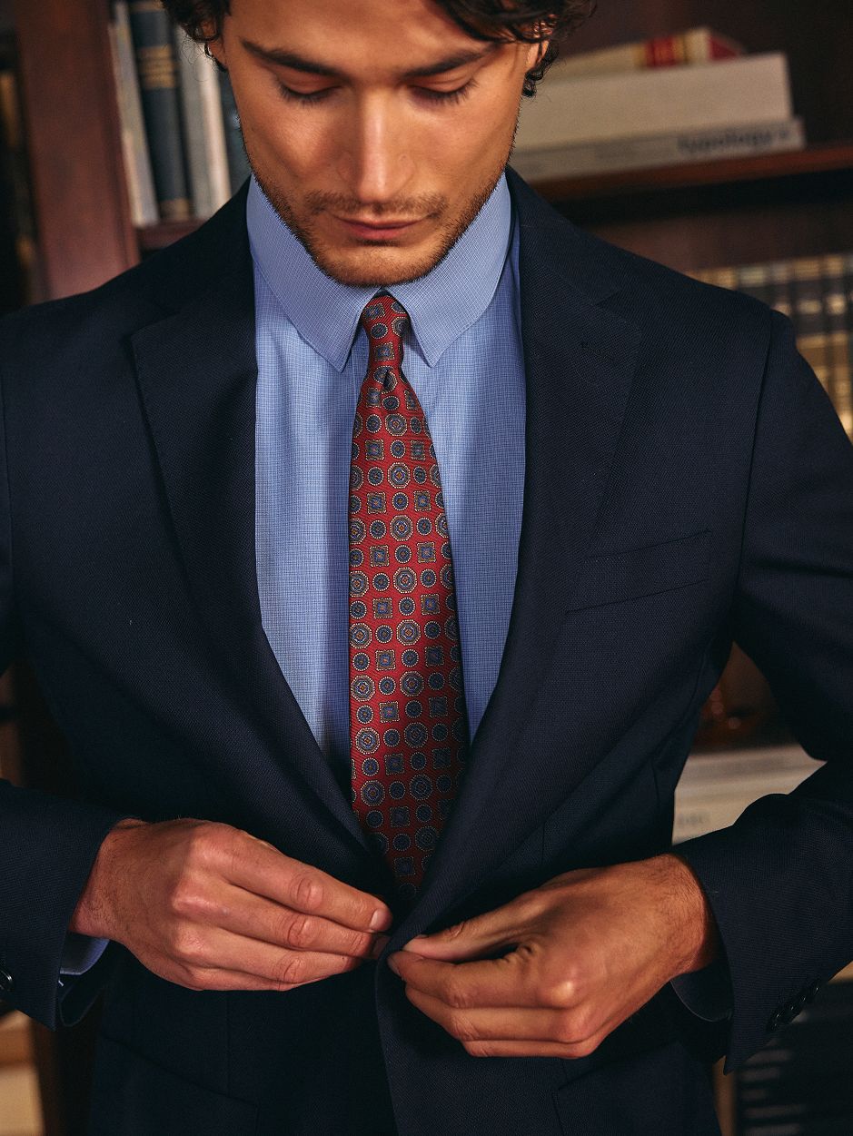 A man looking down while fastening the button on a dark blue suit jacket, paired with a light blue shirt and red patterned tie.
