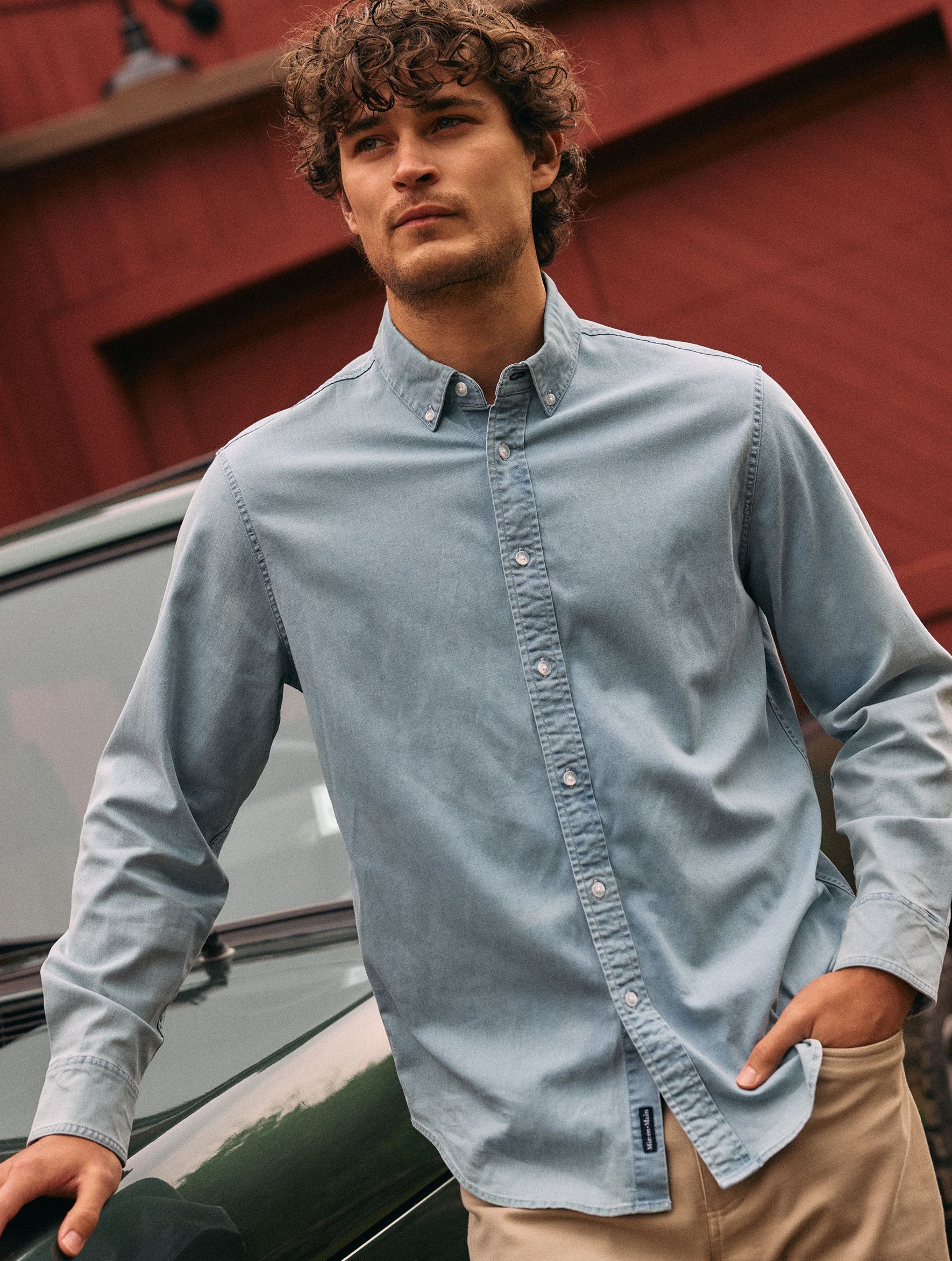 A man with curly hair leaning against a green vehicle in front of a red building, wearing a light blue denim-look button-down shirt and beige pants.