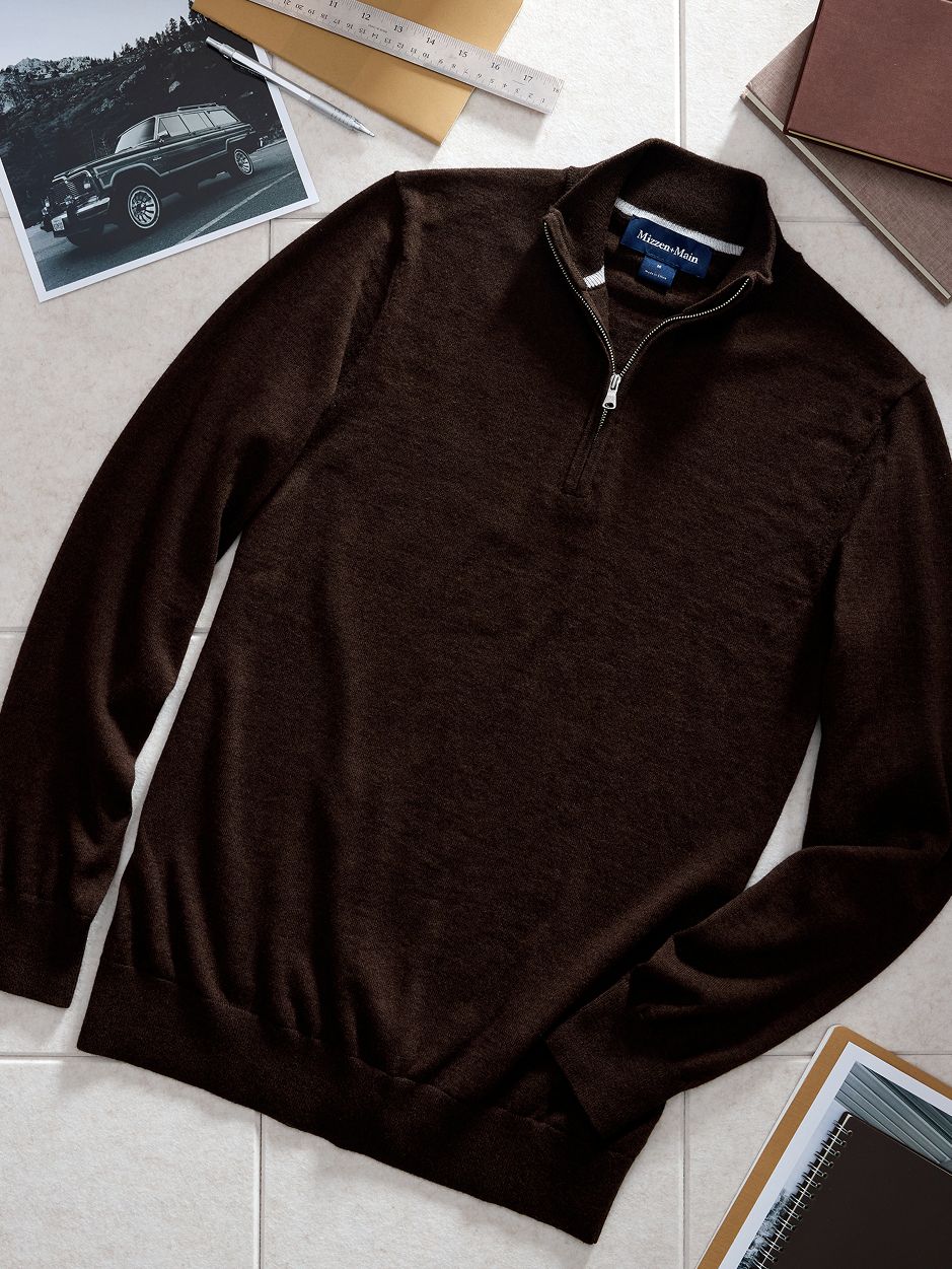 A top-down product flat-lay photo of a dark brown quarter-zip sweater set among office and design props on a tiled surface.