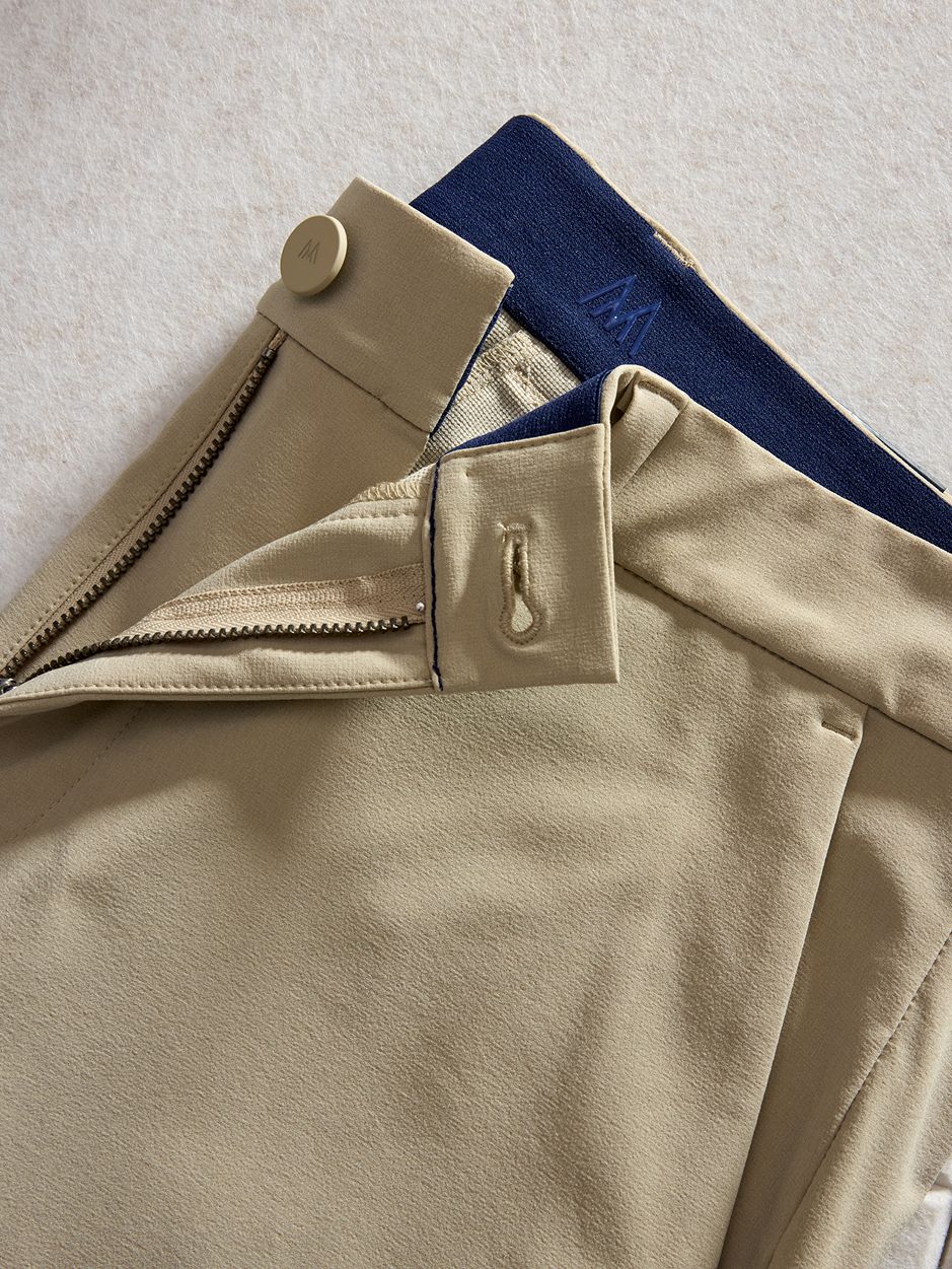 Detail shot of tan trousers showing the open zipper fly, button closure, and interior navy elastic waistband with brand detailing.