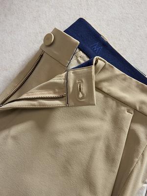 Detail shot of tan trousers showing the open zipper fly, button closure, and interior navy elastic waistband with brand detailing.