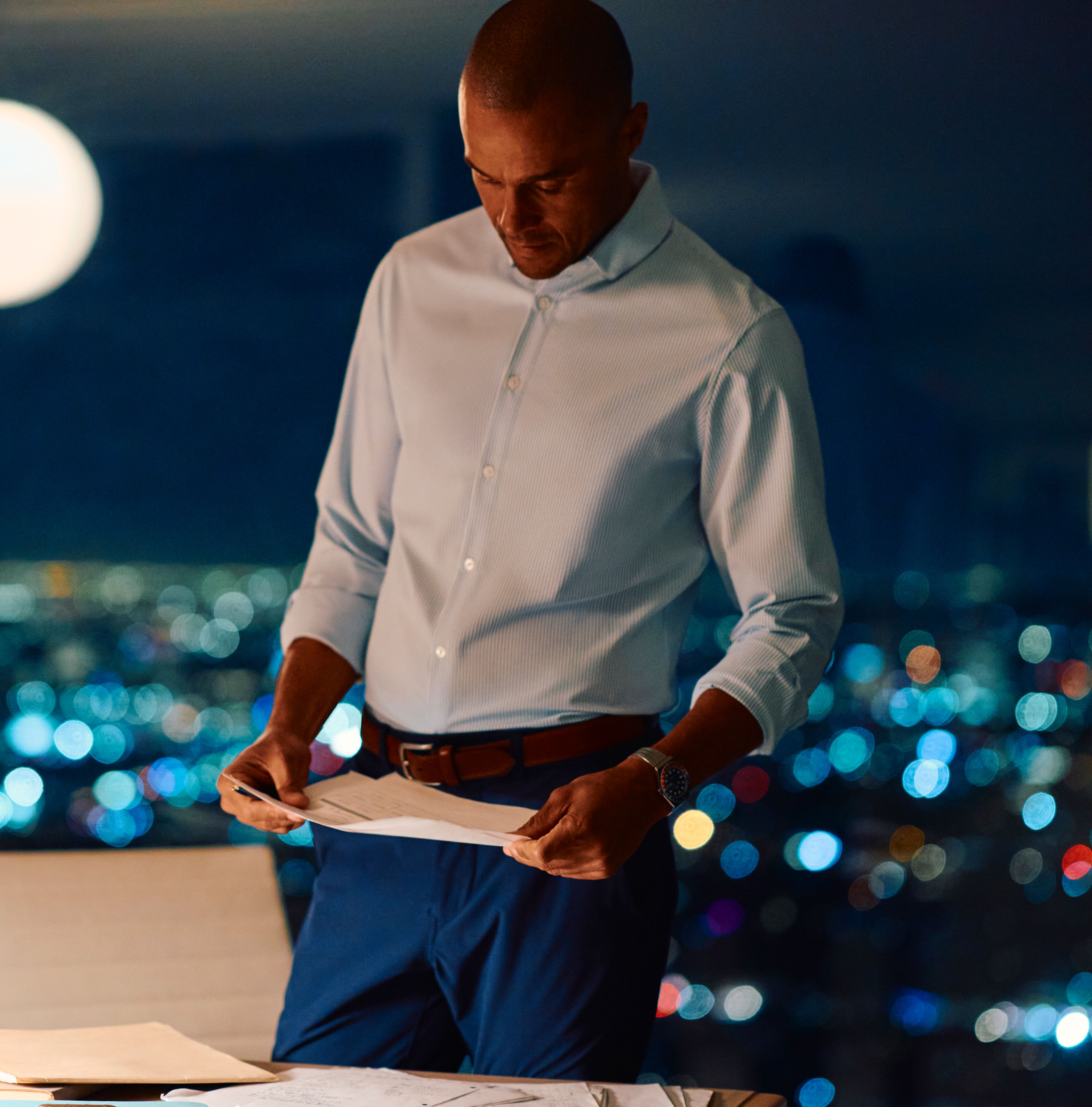 A man reviewing paperwork in a modern high-rise office at night, wearing a light blue dress shirt and blue trousers against a blurred city skyline.