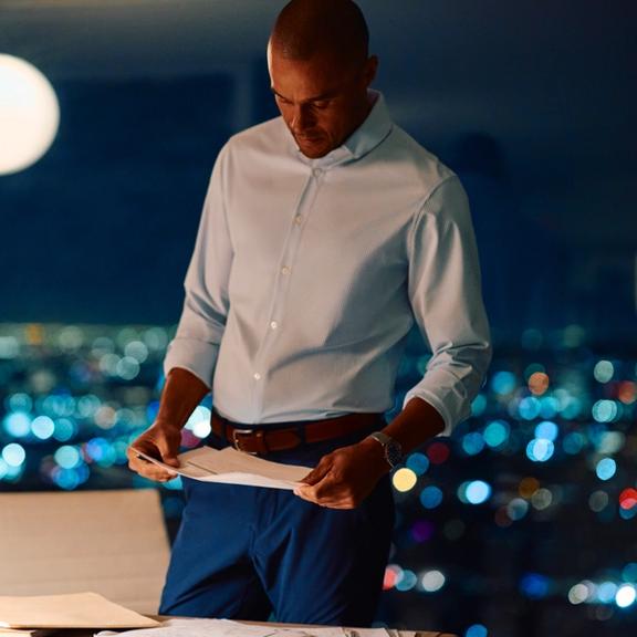 A man reviewing paperwork in a modern high-rise office at night, wearing a light blue dress shirt and blue trousers against a blurred city skyline.