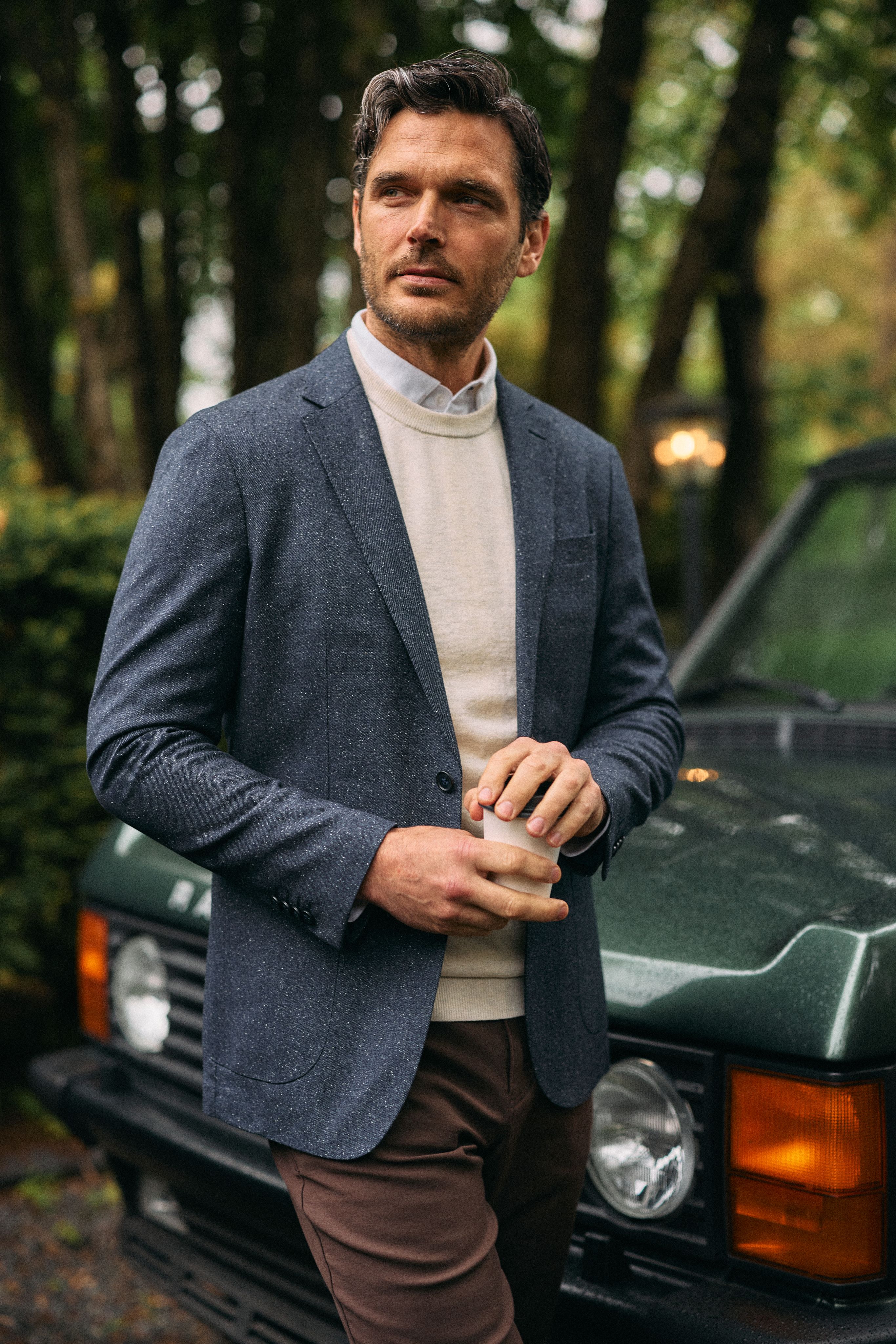 Man in a gray blazer over a cream sweater and collared shirt, holding a glass beside a green Range Rover