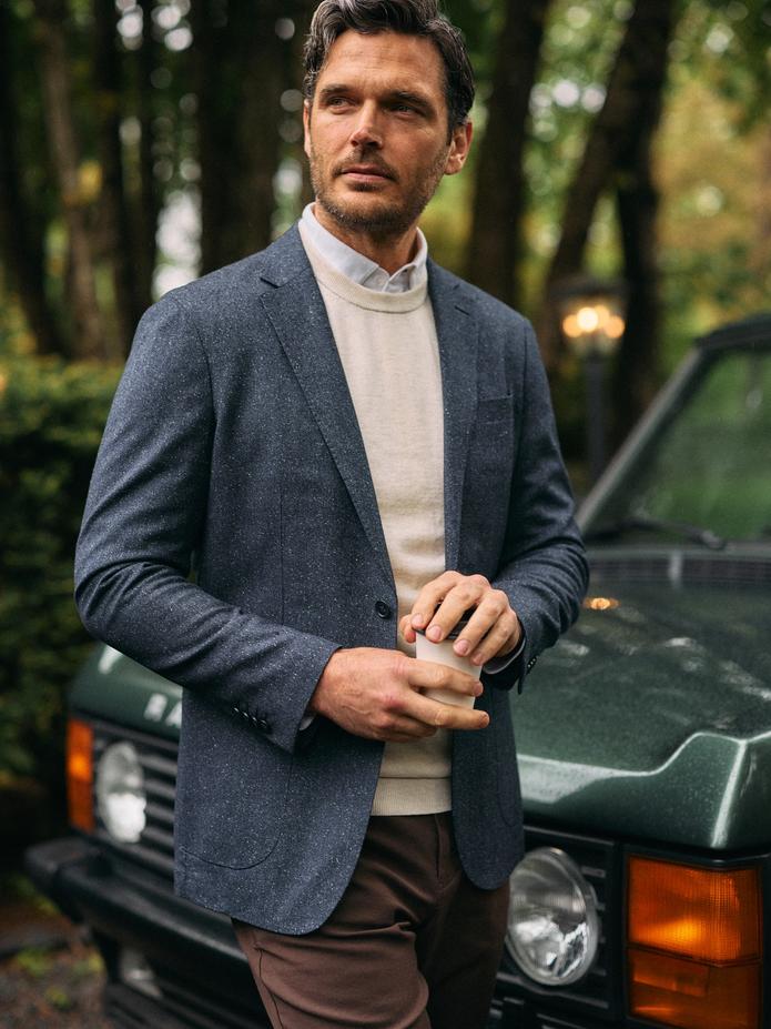 Man in a gray blazer over a cream sweater and collared shirt, holding a glass beside a green Range Rover