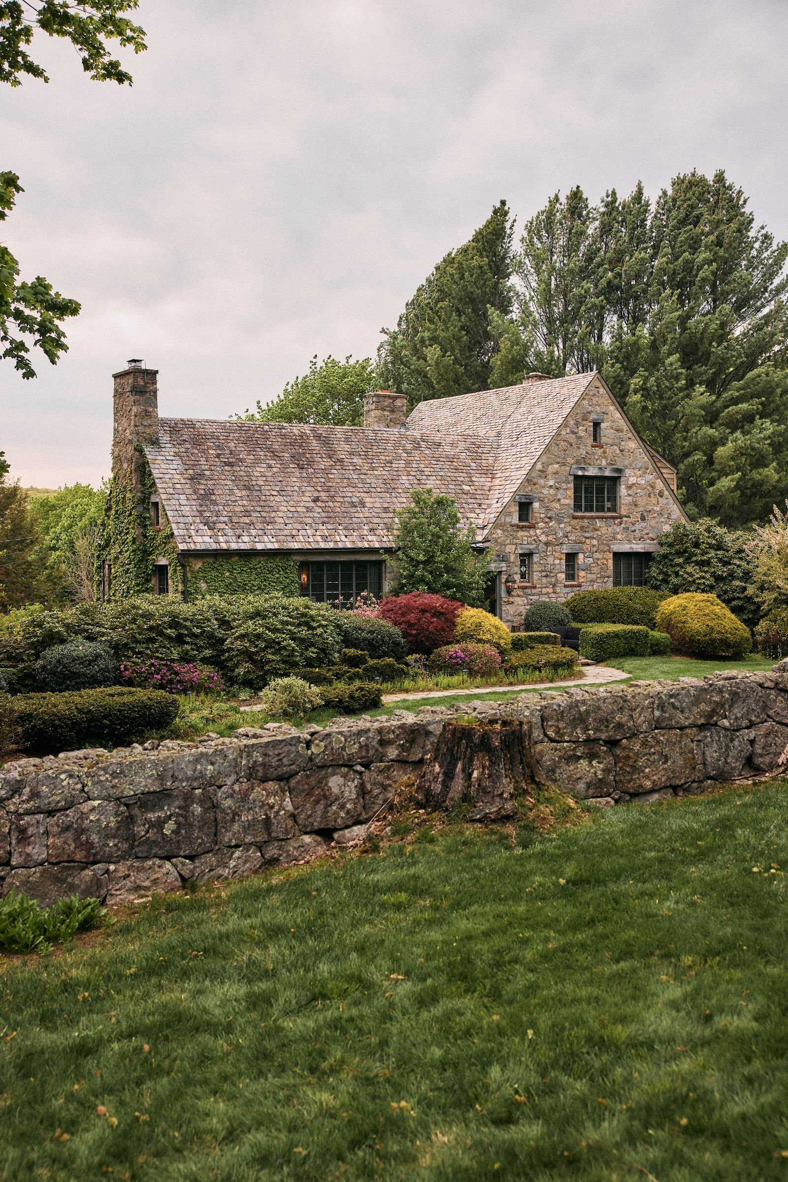 An ivy-covered stone country house in Connecticut behind a low stone garden wall, surrounded by lush gardens and overhanging branches.
