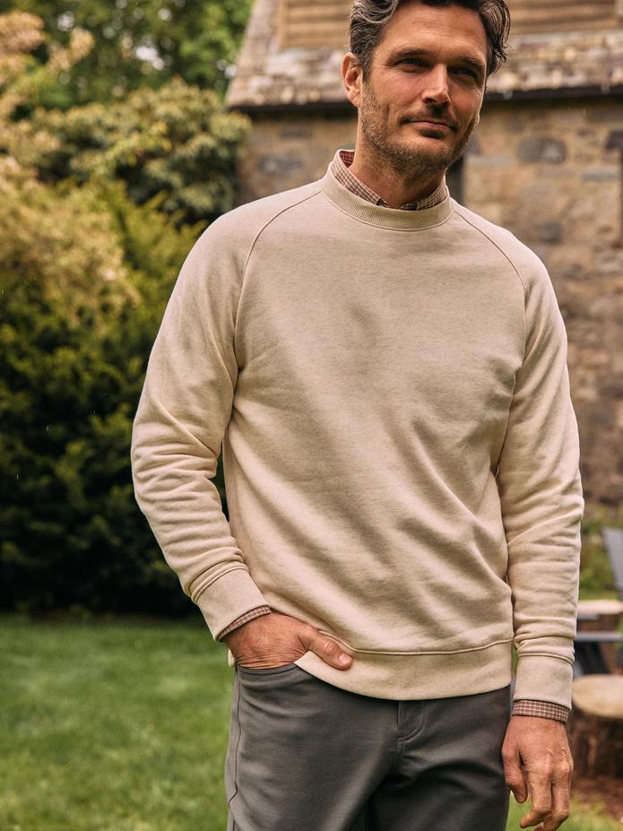 A man wearing a beige crewneck sweatshirt over a plaid collared shirt and grey trousers standing in a garden setting.