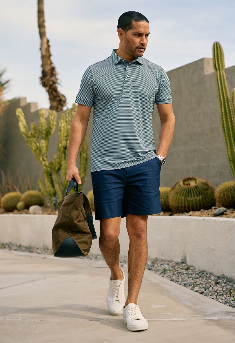 Man in a striped polo and navy Prescott shorts carrying a duffel past desert landscaping