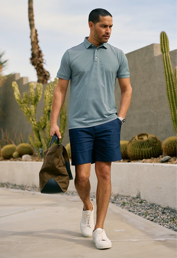 Man in a striped polo and navy Prescott shorts carrying a duffel past desert landscaping