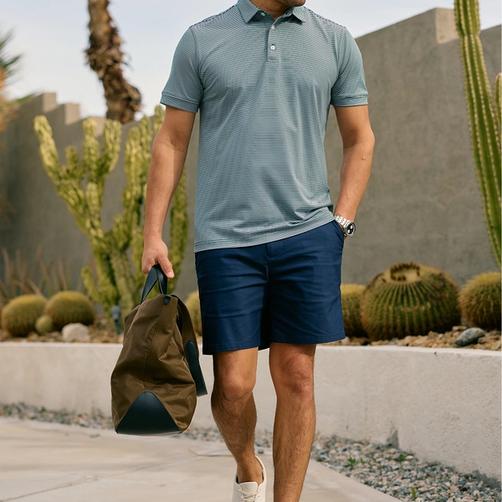 Man in a striped polo and navy Prescott shorts carrying a duffel past desert landscaping