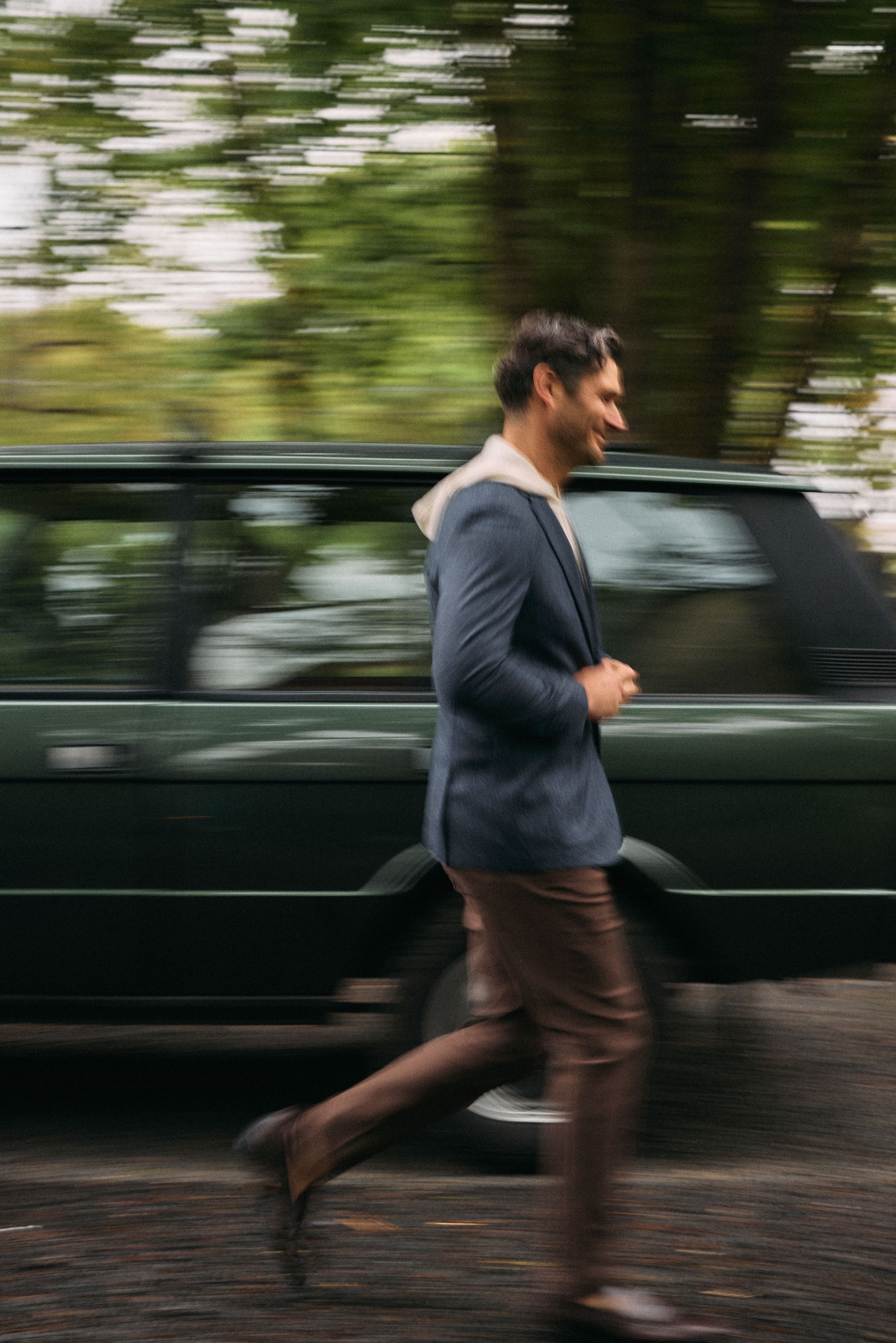 Man in a navy blazer layered over a cream hoodie with brown pants, striding past a green Range Rover